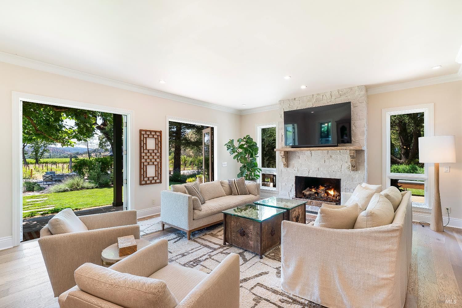 This bright and airy living room features a neutral color palette, plush cream-colored sofas, and a large stone fireplace with a mounted television. The space is anchored by a patterned area rug and two glass-topped coffee tables, while expansive glass doors open to a lush, sunlit vineyard view. The overall aesthetic is sophisticated and inviting, blending comfortable luxury with seamless indoor-outdoor living.