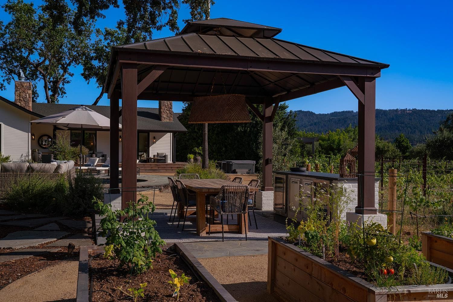 This inviting outdoor living space features a large, covered gazebo housing a wooden dining set and a built-in outdoor kitchen area. The patio is surrounded by raised garden beds and overlooks a scenic landscape with rolling hills in the distance. The setting offers a perfect blend of rustic charm and modern convenience, ideal for al fresco dining and entertaining.