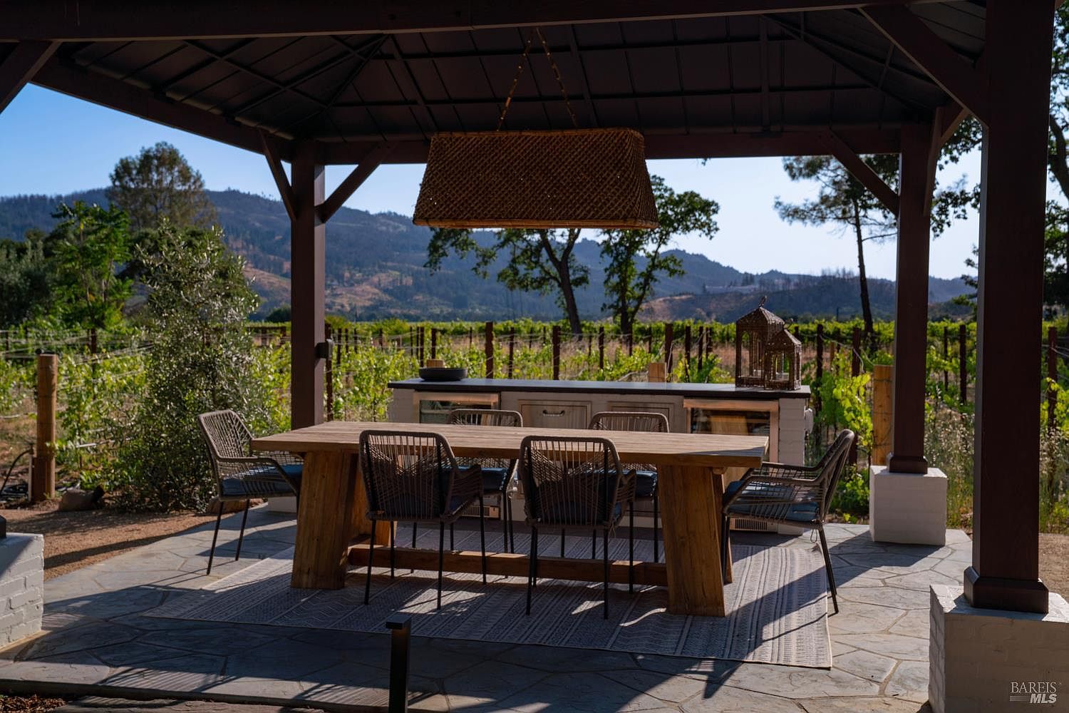 This inviting outdoor dining area is situated under a covered pavilion, featuring a rustic wooden table surrounded by modern woven chairs on a patterned area rug. The space includes a built-in outdoor kitchen counter and offers a stunning, unobstructed view of a lush vineyard set against rolling hills. The setting provides a serene and sophisticated atmosphere, perfect for al fresco dining and entertaining.