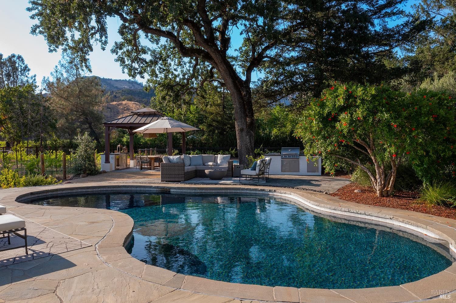 This inviting outdoor oasis features a custom-shaped swimming pool with stone coping, set against a backdrop of mature oak trees and rolling hills. The patio area includes a comfortable lounge seating arrangement, a fire pit, and a built-in outdoor kitchen, all shaded by a large tree and a nearby gazebo. The scene captures a serene, resort-like atmosphere perfect for entertaining and relaxation.