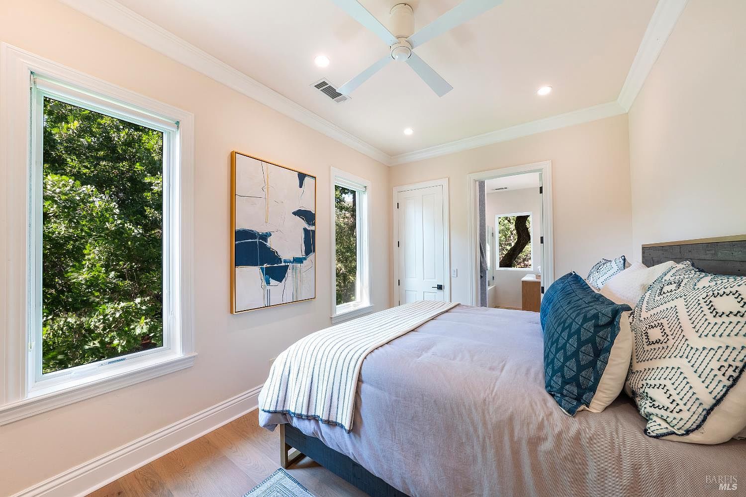 This bright and airy primary bedroom features light-toned walls, hardwood flooring, and a large window that brings in abundant natural light. The room is furnished with a comfortable bed adorned with patterned blue and white pillows, complemented by a modern abstract art piece on the wall. A ceiling fan and recessed lighting provide a clean, contemporary aesthetic, while a doorway leads into an adjoining bathroom.
