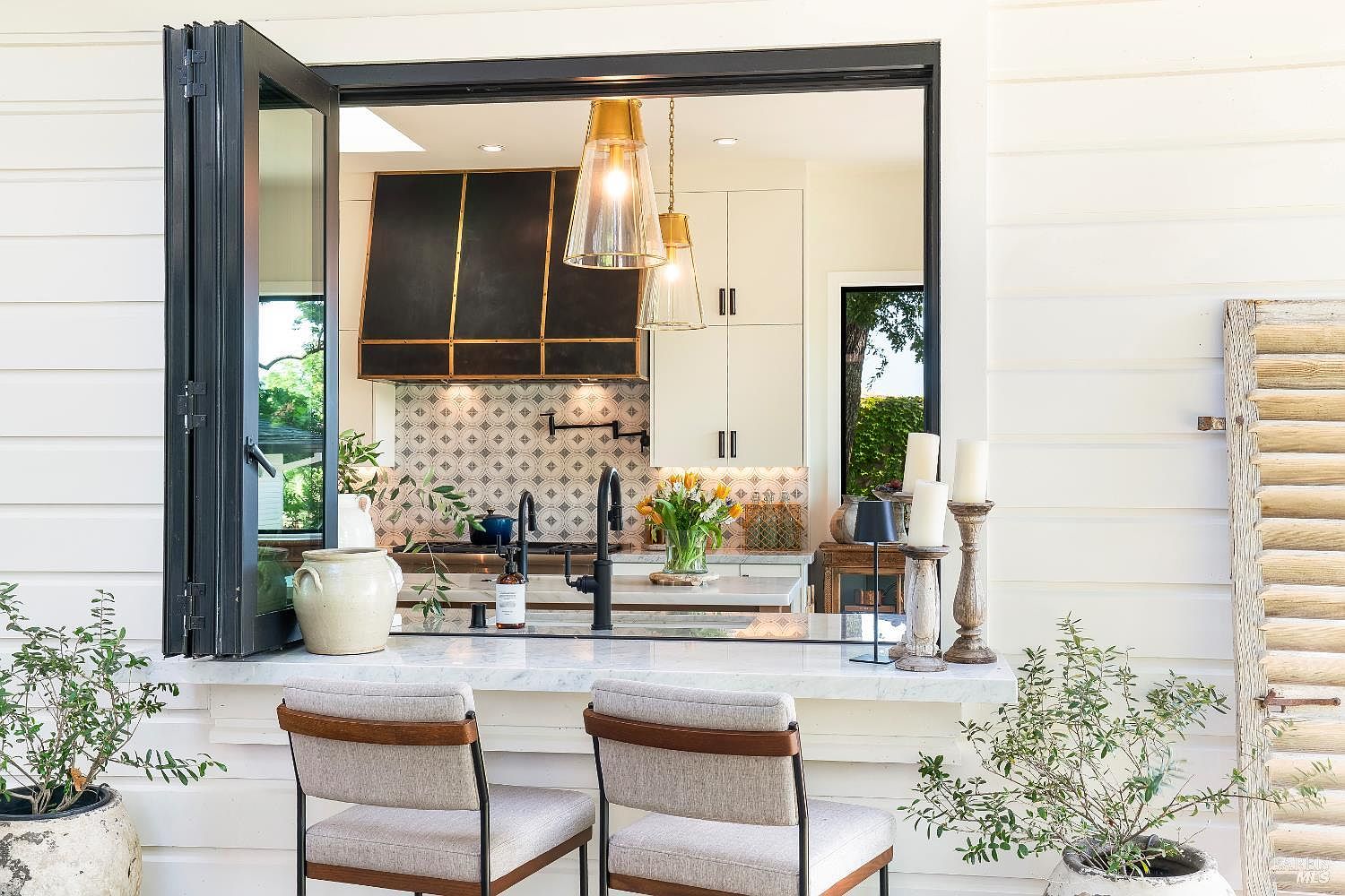 This image captures a seamless indoor-outdoor transition, featuring a pass-through window with folding doors that connects a patio seating area to a stylish kitchen. The outdoor space includes two modern upholstered bar stools set against a marble countertop, while the interior reveals a sophisticated kitchen with a dark metal range hood and patterned backsplash. The perspective is eye-level, emphasizing the inviting, open-concept design that blends luxury indoor living with a relaxed outdoor atmosphere.