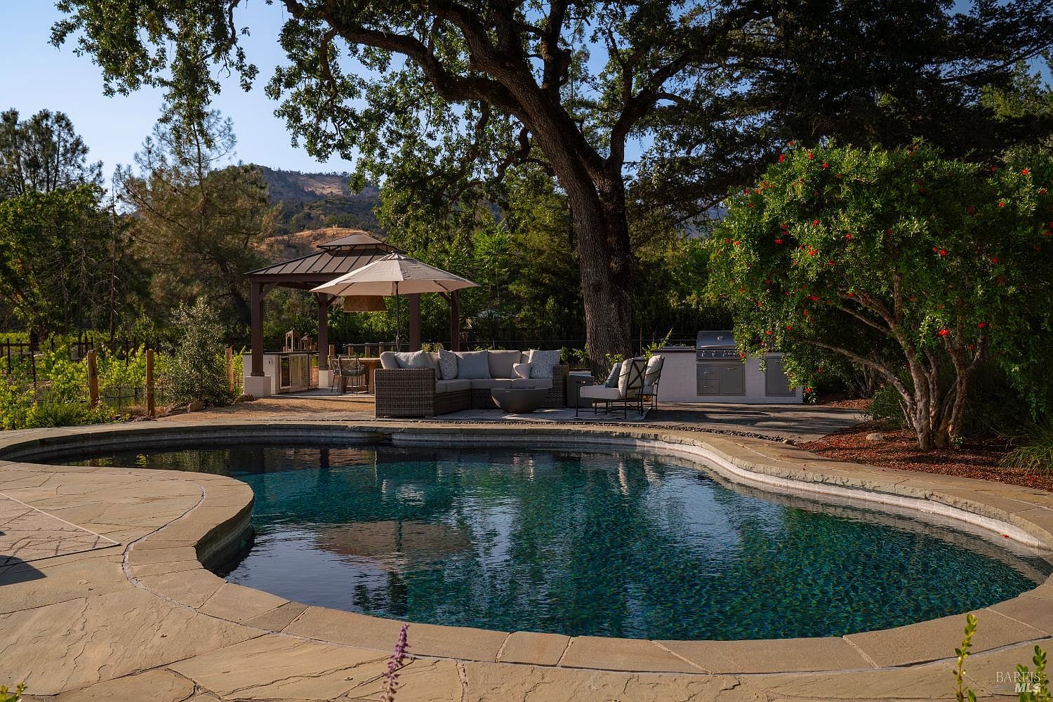 This inviting outdoor living space features a custom-shaped swimming pool with natural stone coping, set against a backdrop of mature oak trees and rolling hills. The patio area includes a comfortable sectional sofa, a fire pit, and a covered outdoor kitchen with a gazebo, creating a perfect environment for relaxation and entertaining. The scene is captured from a low, eye-level perspective that emphasizes the seamless transition between the pool deck and the lush, landscaped backyard.