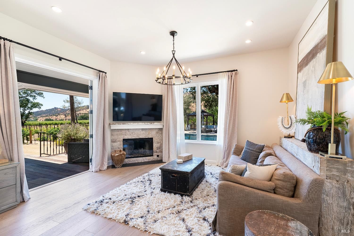 This bright and inviting living room features a cozy fireplace with a stone surround, a plush area rug, and a comfortable sofa, creating a warm atmosphere. Large glass doors open directly onto a deck, seamlessly blending indoor and outdoor living while offering scenic views of the surrounding vineyard. The space is elegantly styled with a rustic-chic aesthetic, highlighted by a statement chandelier and tasteful decor.