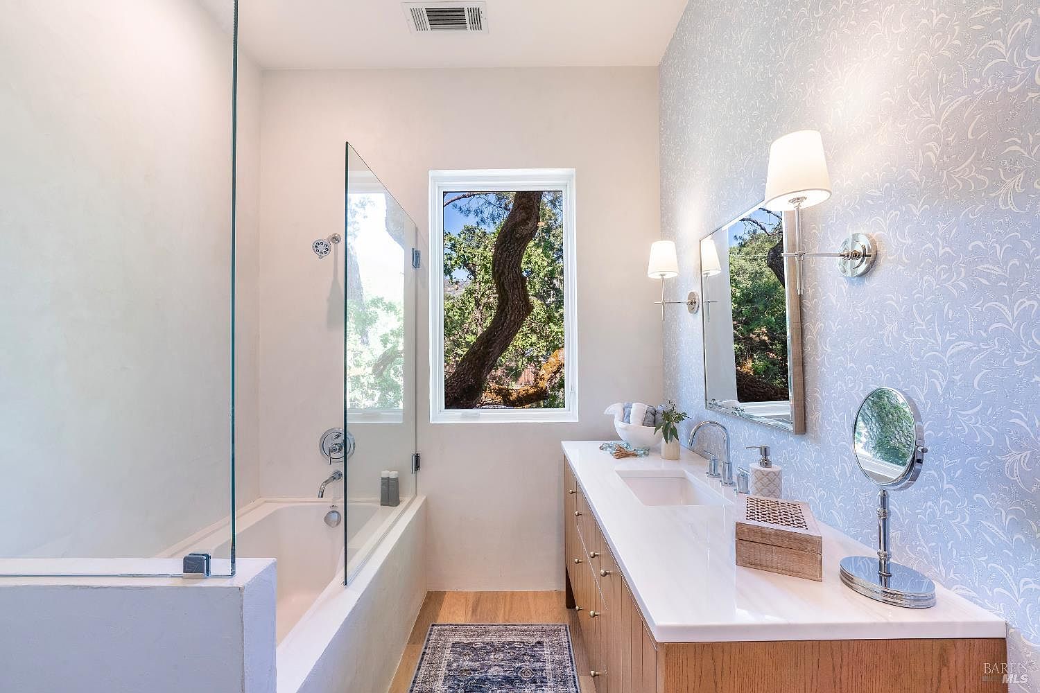 This elegant primary bathroom features a sleek, modern vanity with a white countertop, dual sinks, and light wood cabinetry, complemented by a striking blue and white patterned wallpaper. A glass-enclosed shower and bathtub combination sits adjacent to a large window that frames a serene, tree-filled outdoor view. The space is bright and airy, enhanced by sophisticated wall-mounted lighting fixtures and a minimalist aesthetic.