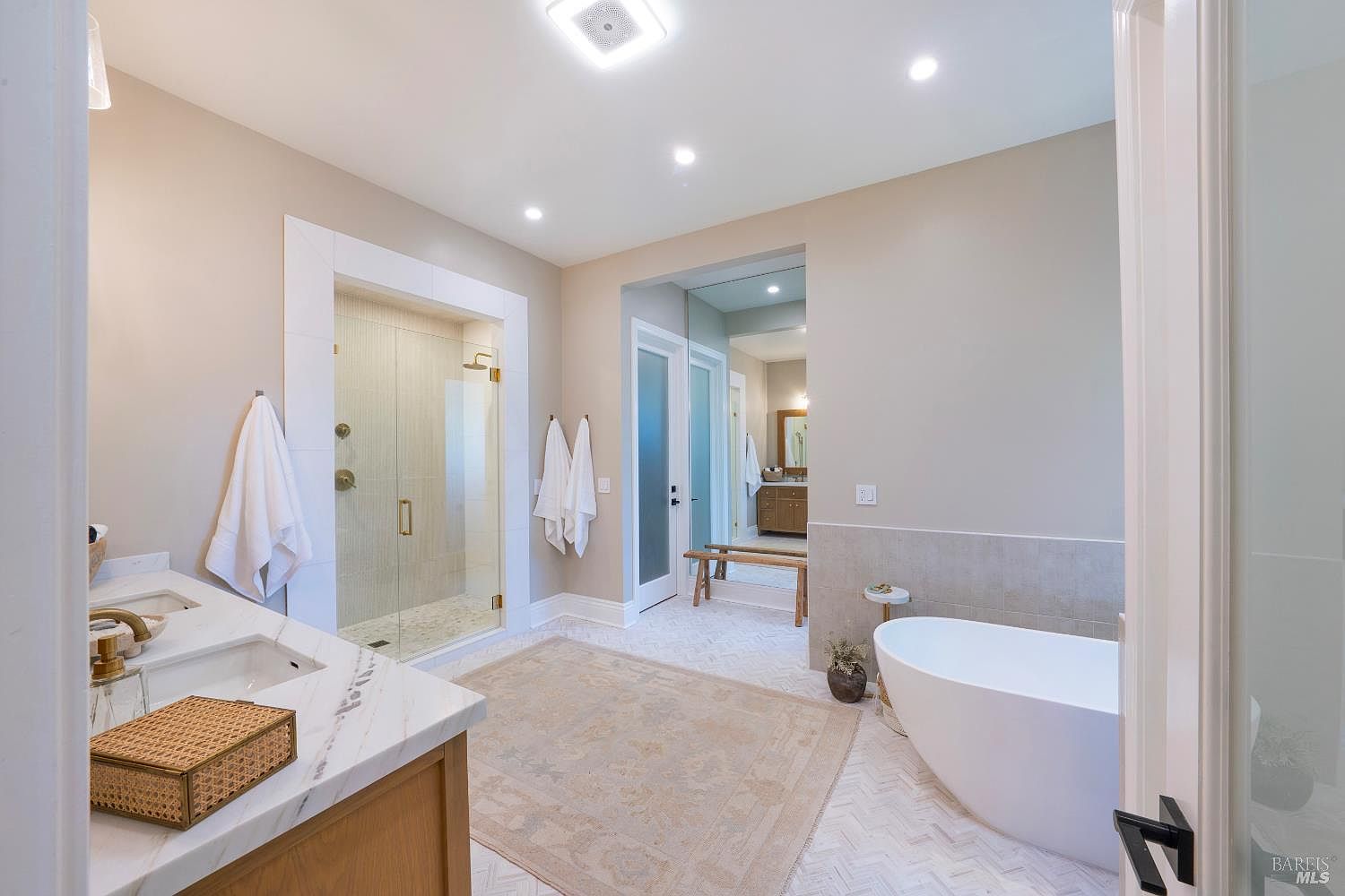 This luxurious primary bathroom features a clean, modern aesthetic with a double vanity, a spacious glass-enclosed walk-in shower, and a sleek freestanding soaking tub. The room is accented by a herringbone-patterned tile floor, a large area rug, and warm, neutral wall tones that create a serene and spa-like atmosphere. The perspective is from the doorway, looking into the bright, well-lit space that flows seamlessly into an adjacent dressing area.
