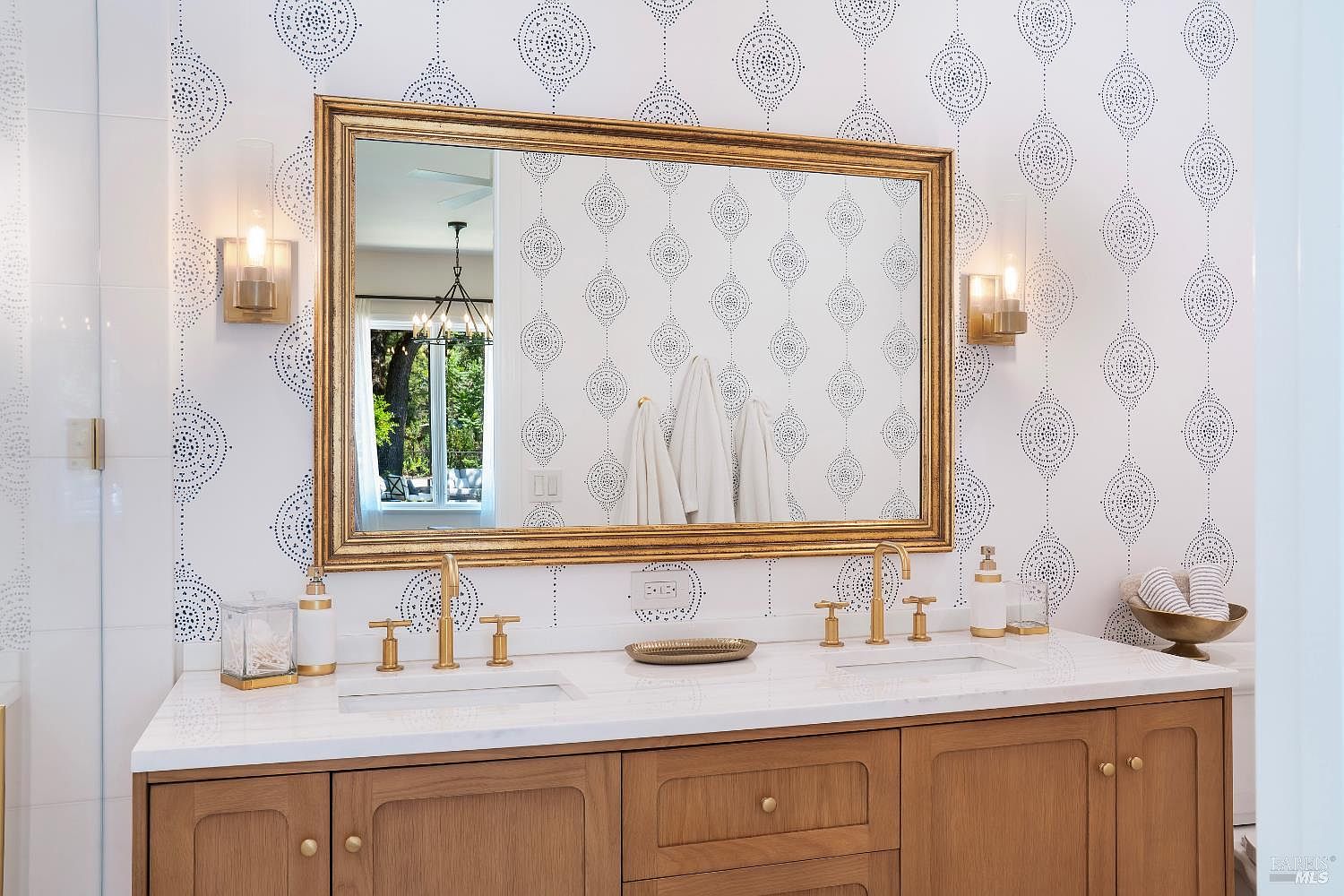 This elegant primary bathroom features a double vanity with a light wood finish, topped with a clean white countertop and gold-toned fixtures. A large, ornate gold-framed mirror hangs above the sinks, centered against a striking white wallpaper with a blue geometric pattern. The space is illuminated by two sleek, modern wall sconces, creating a bright and sophisticated atmosphere.