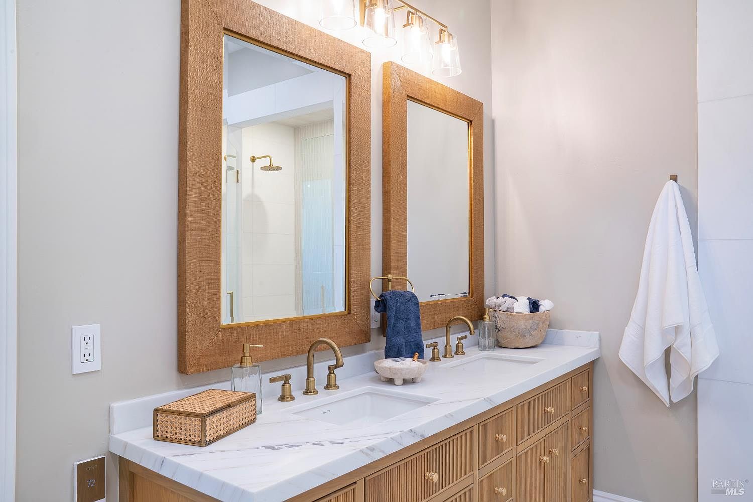 This elegant primary bathroom features a double vanity with a white marble countertop, light wood cabinetry with vertical detailing, and two large mirrors framed in a textured, natural material. The space is accented with warm brass fixtures, including widespread faucets and a multi-light vanity fixture, creating a sophisticated and spa-like atmosphere. A clean, neutral color palette and high-end finishes contribute to a bright and inviting aesthetic.