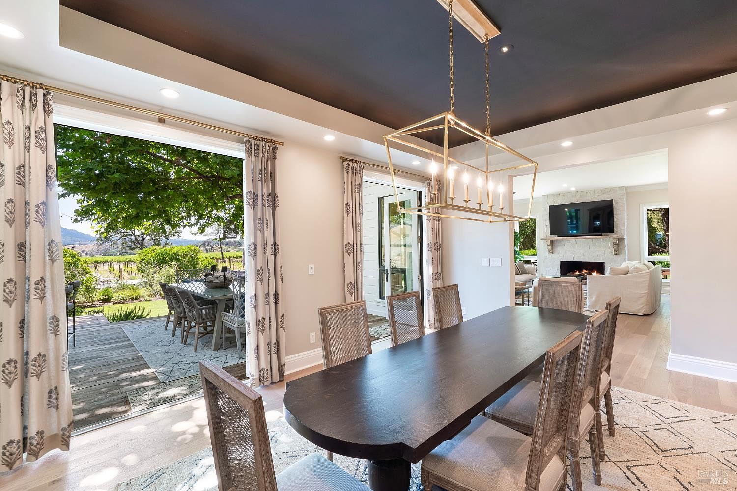 This elegant dining room features a long, dark wood table surrounded by light-toned, cane-back chairs, illuminated by a modern gold geometric chandelier. The space flows seamlessly into an outdoor patio through large sliding glass doors, offering a picturesque view of a lush vineyard and garden. A glimpse into the adjacent living room reveals a cozy fireplace and a neutral color palette, creating a sophisticated and inviting atmosphere.