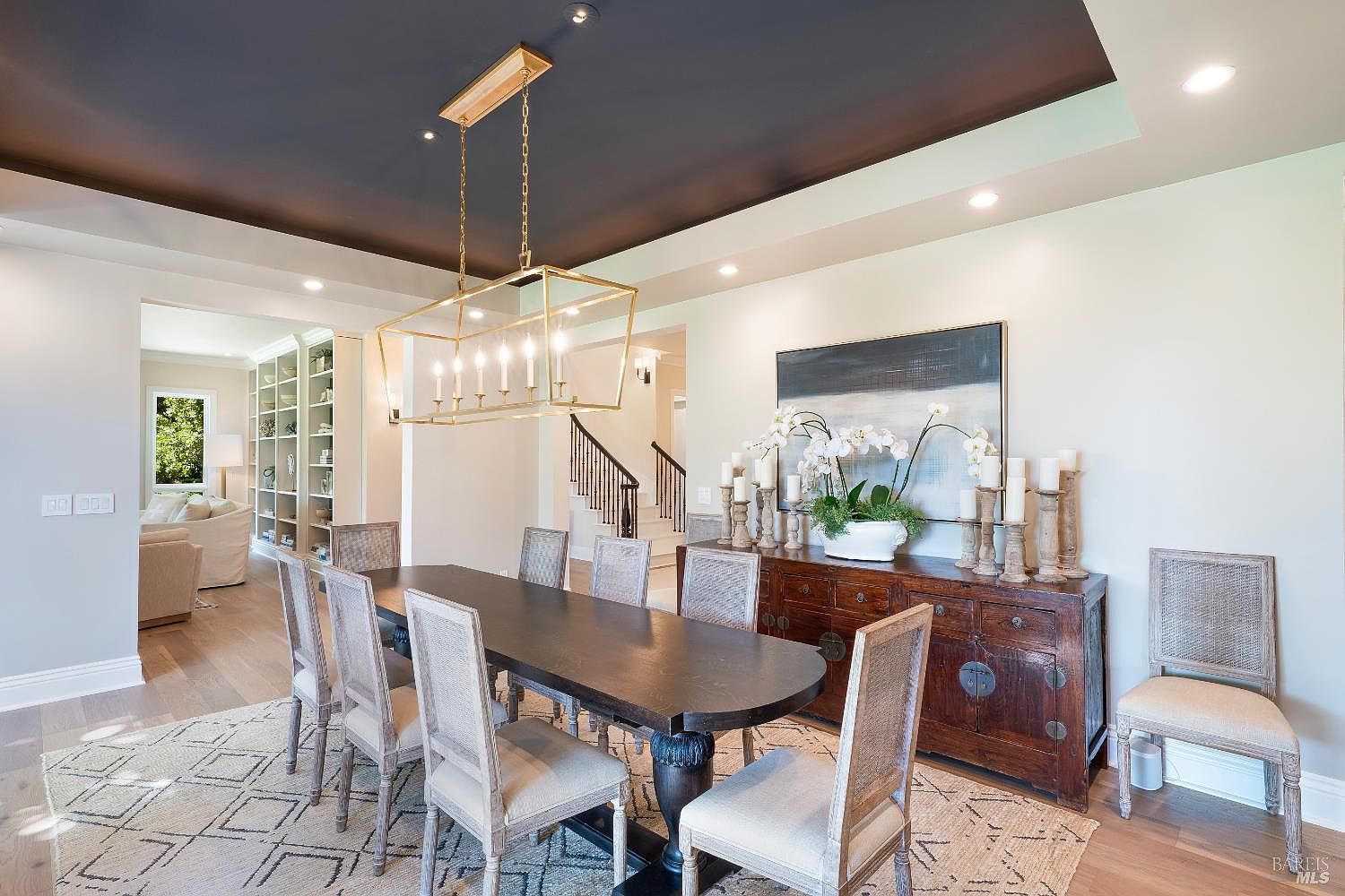 This elegant dining room features a long, dark wood table surrounded by upholstered chairs with cane backs, set beneath a sophisticated gold geometric chandelier. A rich, dark-stained wooden sideboard adorned with candles and a floral arrangement sits against the wall, while a patterned area rug anchors the space. The room is framed by a dark ceiling accent and offers a glimpse into a bright, open-concept living area and a staircase in the background, creating a refined and inviting atmosphere.