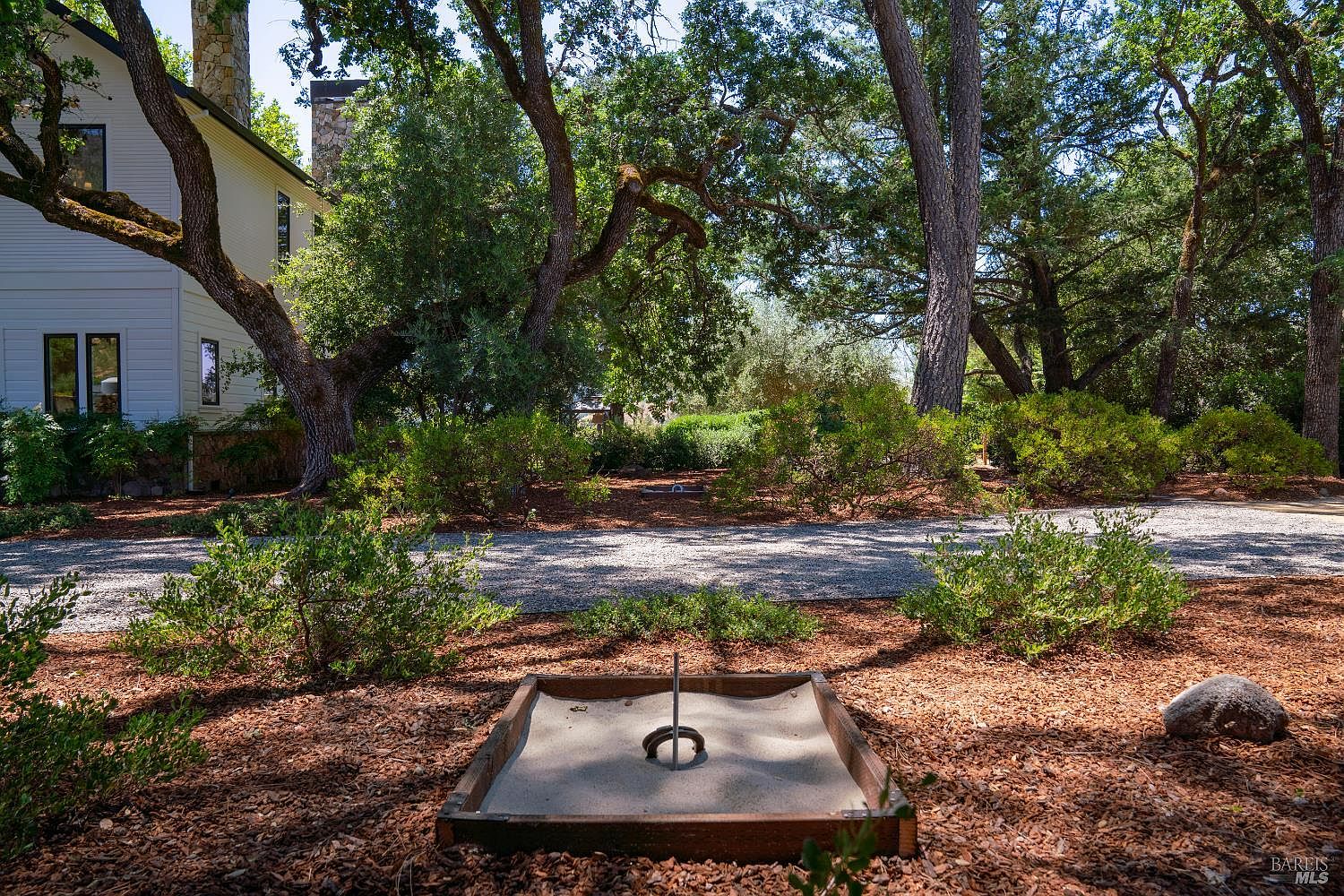 This outdoor scene features a horseshoe pit set in a sandy box, positioned in the foreground of a spacious, wooded yard. The property includes a white-sided house with stone accents visible to the left, surrounded by mature oak trees and natural landscaping. The perspective is a low-angle shot looking across the gravel driveway toward the house, creating a serene and recreational atmosphere.