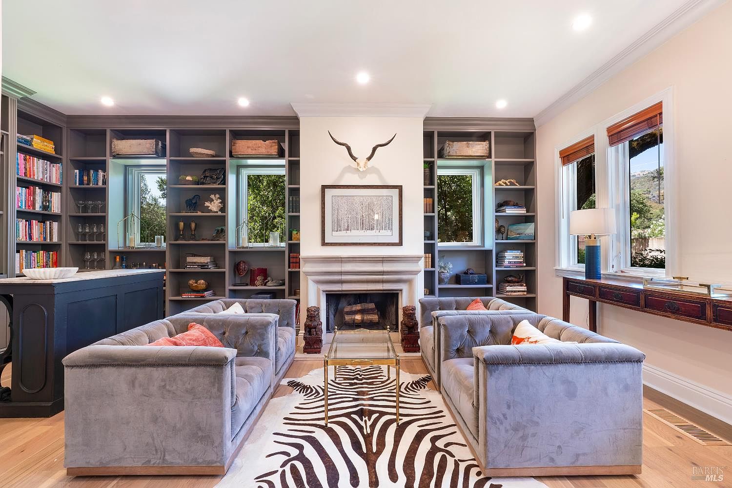 This sophisticated living room features a symmetrical layout centered around a grand stone fireplace adorned with a decorative animal skull and framed artwork. The space is framed by extensive built-in grey shelving units that flank the fireplace and windows, creating a library-like atmosphere. Two plush, tufted grey velvet sofas face each other over a striking zebra-print area rug, establishing a luxurious and balanced aesthetic.