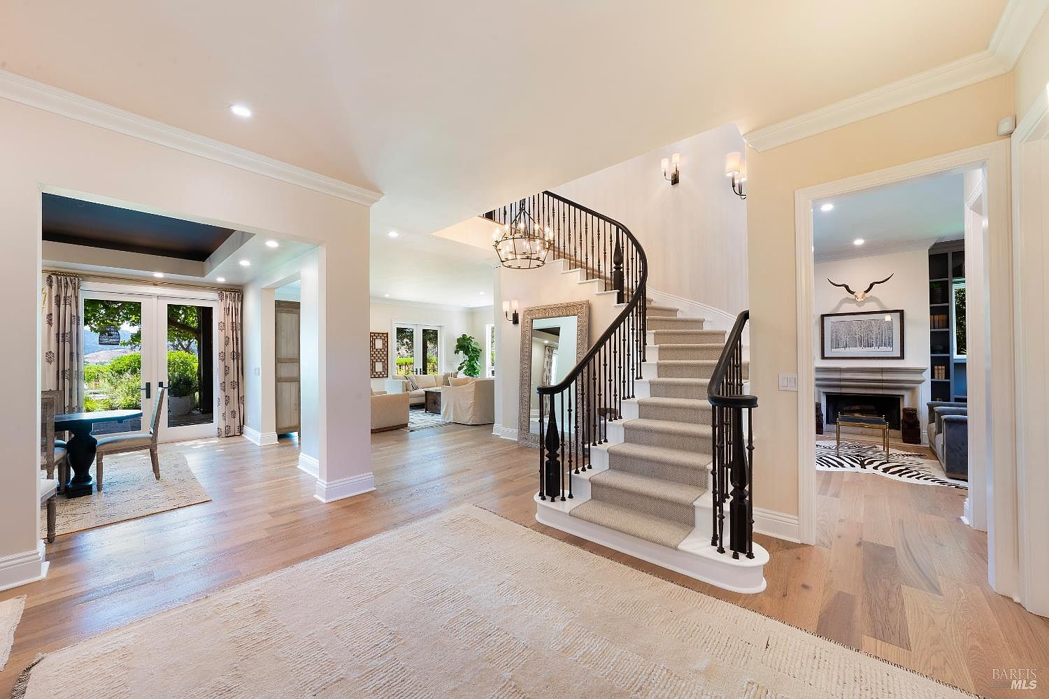 This elegant foyer features a grand, curved staircase with a dark iron railing and a plush carpet runner, serving as the central focal point of the home. To the left, a bright dining area with French doors opens to the outdoors, while a doorway to the right leads into a cozy living room with a fireplace. The space is characterized by light hardwood floors, neutral walls, and sophisticated architectural details, creating a welcoming and luxurious atmosphere.