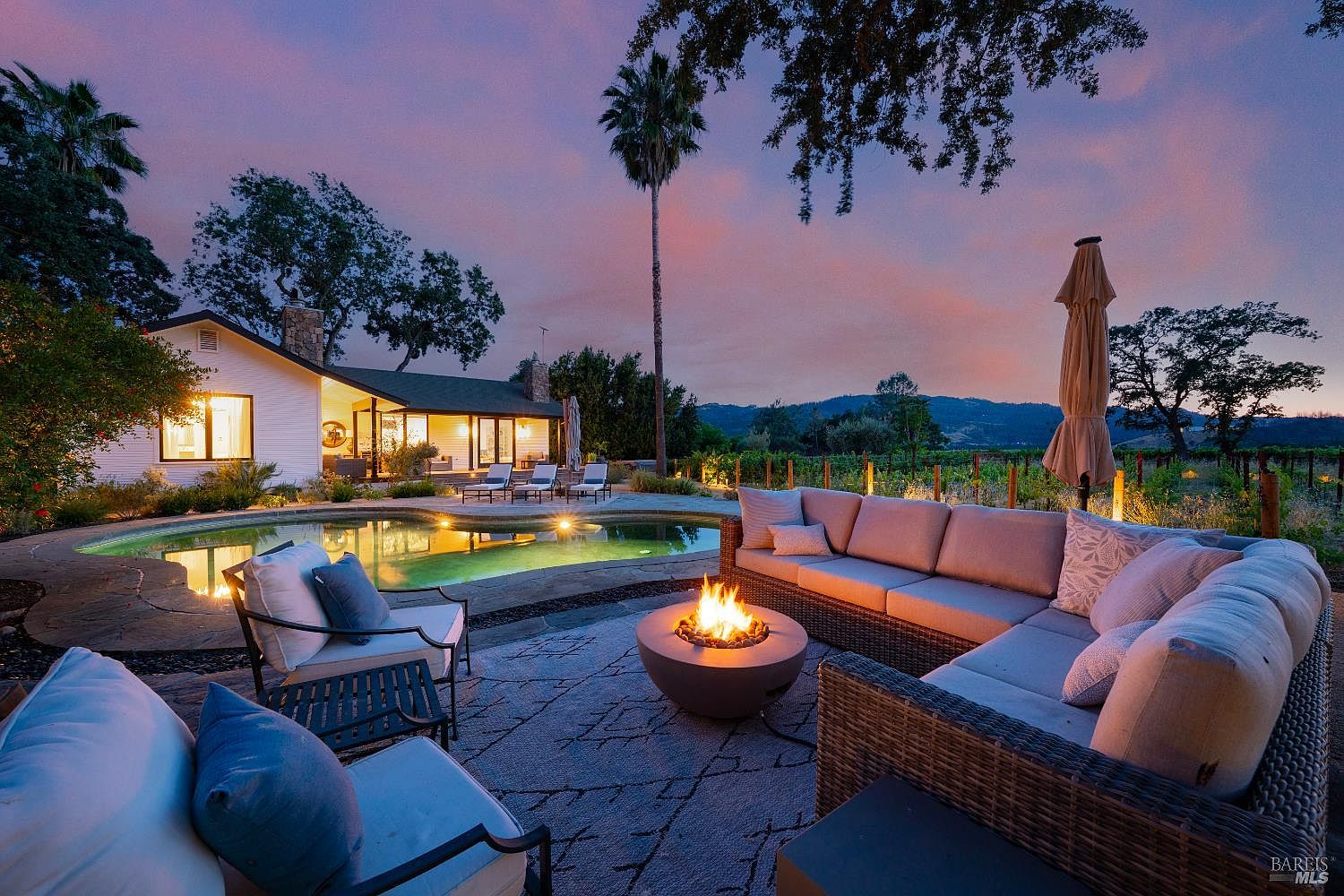 This stunning outdoor living space features a cozy fire pit surrounded by plush, modern wicker seating, perfect for evening relaxation. The area overlooks a beautifully illuminated swimming pool and a serene vineyard landscape under a vibrant sunset sky. The scene captures an inviting, luxurious atmosphere that highlights the home's seamless integration with its picturesque surroundings.