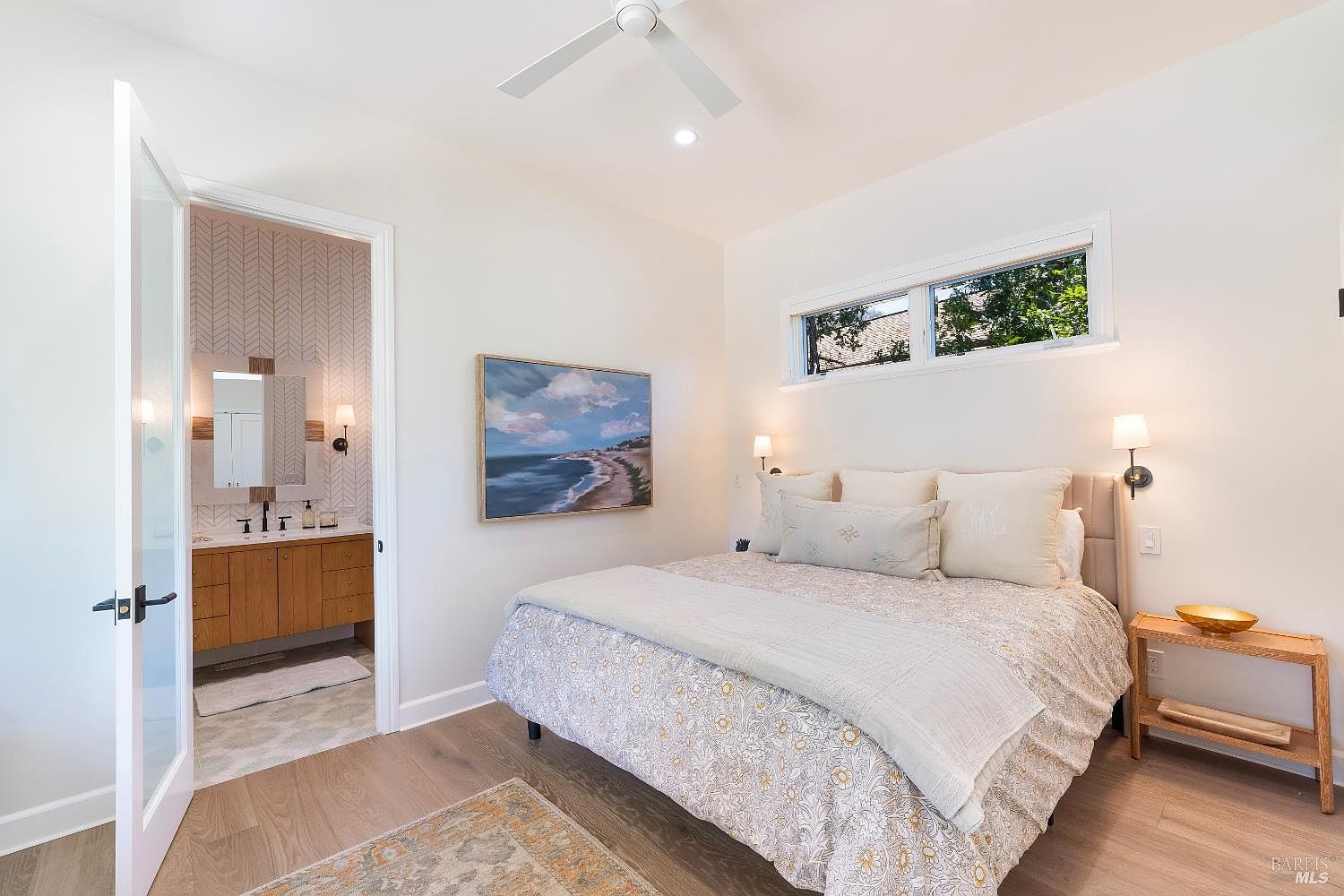 This bright and airy primary bedroom features light wood flooring, a neutral color palette, and a comfortable bed dressed in patterned bedding. A large piece of coastal-themed artwork hangs on the wall, while an open doorway reveals a stylish en-suite bathroom with a herringbone-patterned wall. The room is illuminated by soft wall-mounted sconces and natural light from a horizontal window, creating a serene and inviting atmosphere.