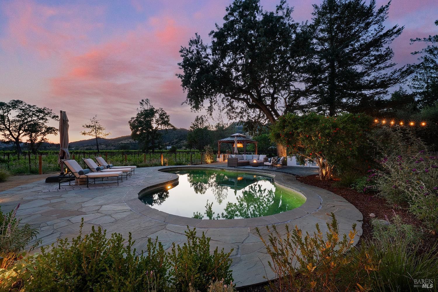 This stunning outdoor oasis features a custom-shaped swimming pool surrounded by natural stone decking, set against a backdrop of rolling hills and a vineyard at sunset. The area includes a cozy lounge space under a wooden gazebo, comfortable poolside loungers, and mature landscaping illuminated by soft string lights. The overall atmosphere is serene and luxurious, perfect for high-end outdoor entertaining.