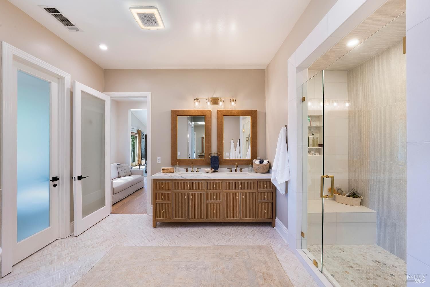 This elegant primary bathroom features a double vanity with a warm wood finish, topped with white stone countertops and framed mirrors. To the right, a spacious walk-in glass shower includes a built-in bench and pebble-tiled flooring, while the room is grounded by a light-colored herringbone tile floor and a soft area rug. The space feels bright and airy, with a neutral color palette and a view into an adjacent living area through a doorway.