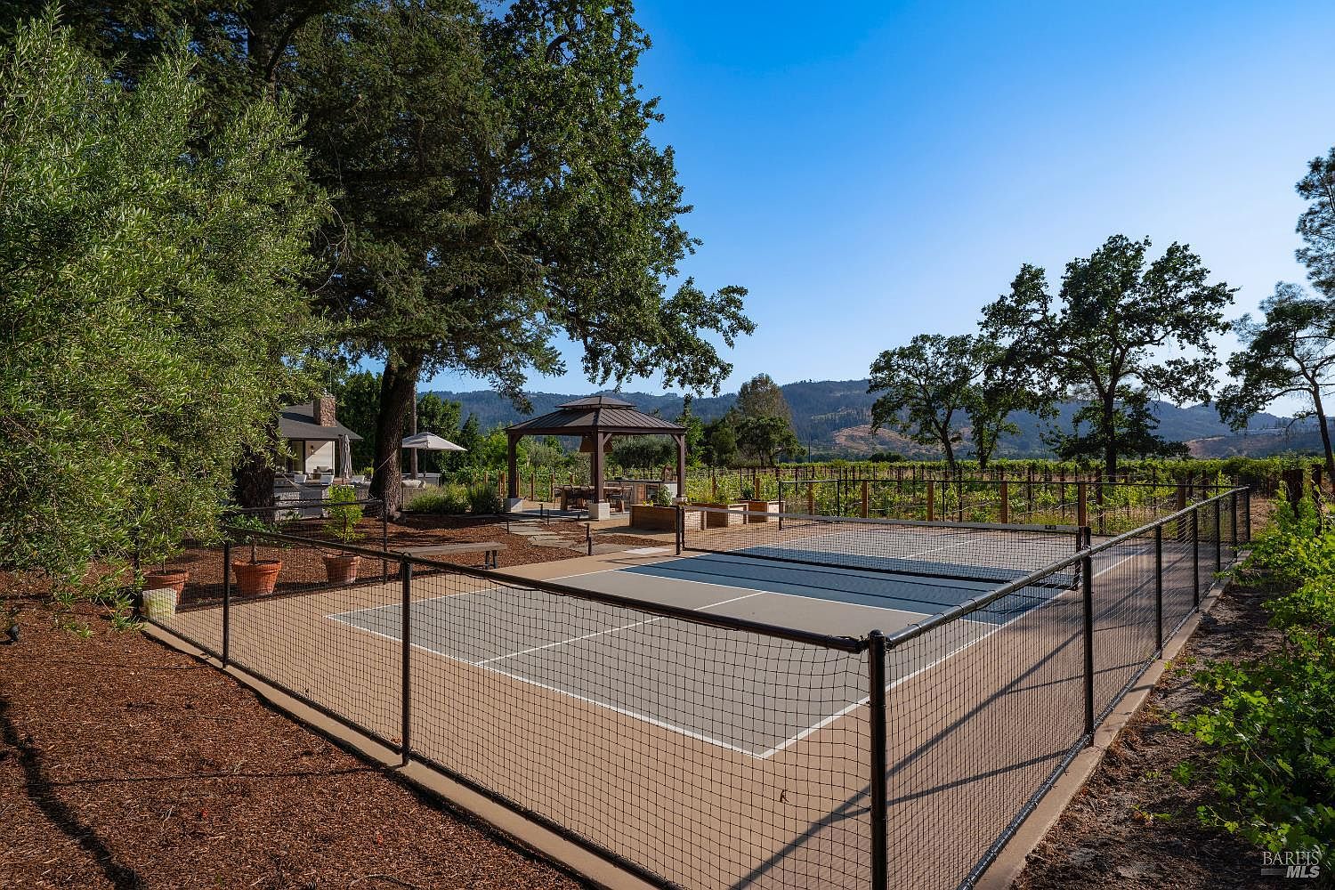 This outdoor space features a well-maintained pickleball court enclosed by black chain-link fencing, set against a backdrop of lush trees and a vineyard. A nearby gazebo provides a shaded seating area, creating an inviting atmosphere for recreation and relaxation in a scenic, rural setting. The perspective captures the court in the foreground, emphasizing the property's lifestyle amenities and expansive natural surroundings.