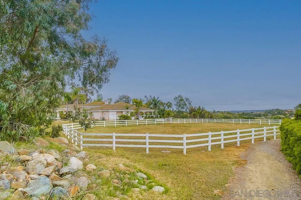 The image showcases a spacious yard with a white, three-rail fence enclosing a grassy area, possibly a horse paddock or large garden. A single-story house is visible in the background, partially obscured by trees, suggesting a rural or suburban setting. A dirt path runs along the edge of the yard, adding to the property's rustic charm.