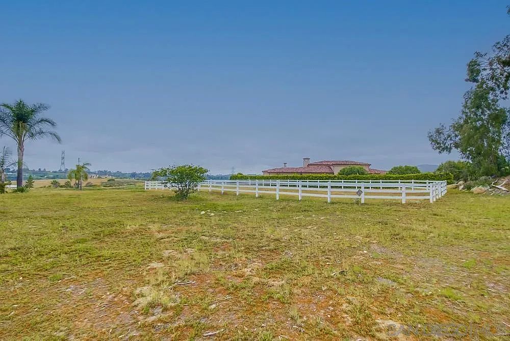 The image showcases a sprawling yard with a well-maintained lawn, partially dry, enclosed by a white fence. In the background, a large house with a red tile roof is visible, adding to the property's appeal. A palm tree stands to the left, and other trees are scattered around the property, enhancing the landscape's natural beauty.