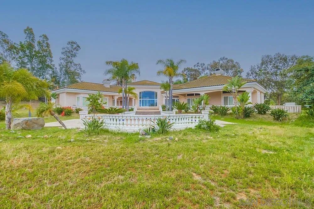 This image showcases the front exterior of a luxurious single-story home with a sprawling lawn. The house features a light-colored facade, a tiled roof, and a decorative white balustrade. Palm trees and other greenery enhance the property's curb appeal, creating a serene and inviting atmosphere.