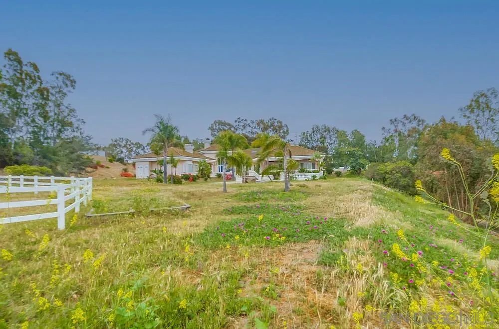 This image showcases a sprawling yard with a well-maintained lawn and vibrant wildflowers in the foreground. A white fence adds a touch of rustic charm, leading the eye towards a large, luxurious house in the background, partially obscured by mature trees and palm trees. The overall impression is one of spaciousness and tranquility, highlighting the property's outdoor appeal.