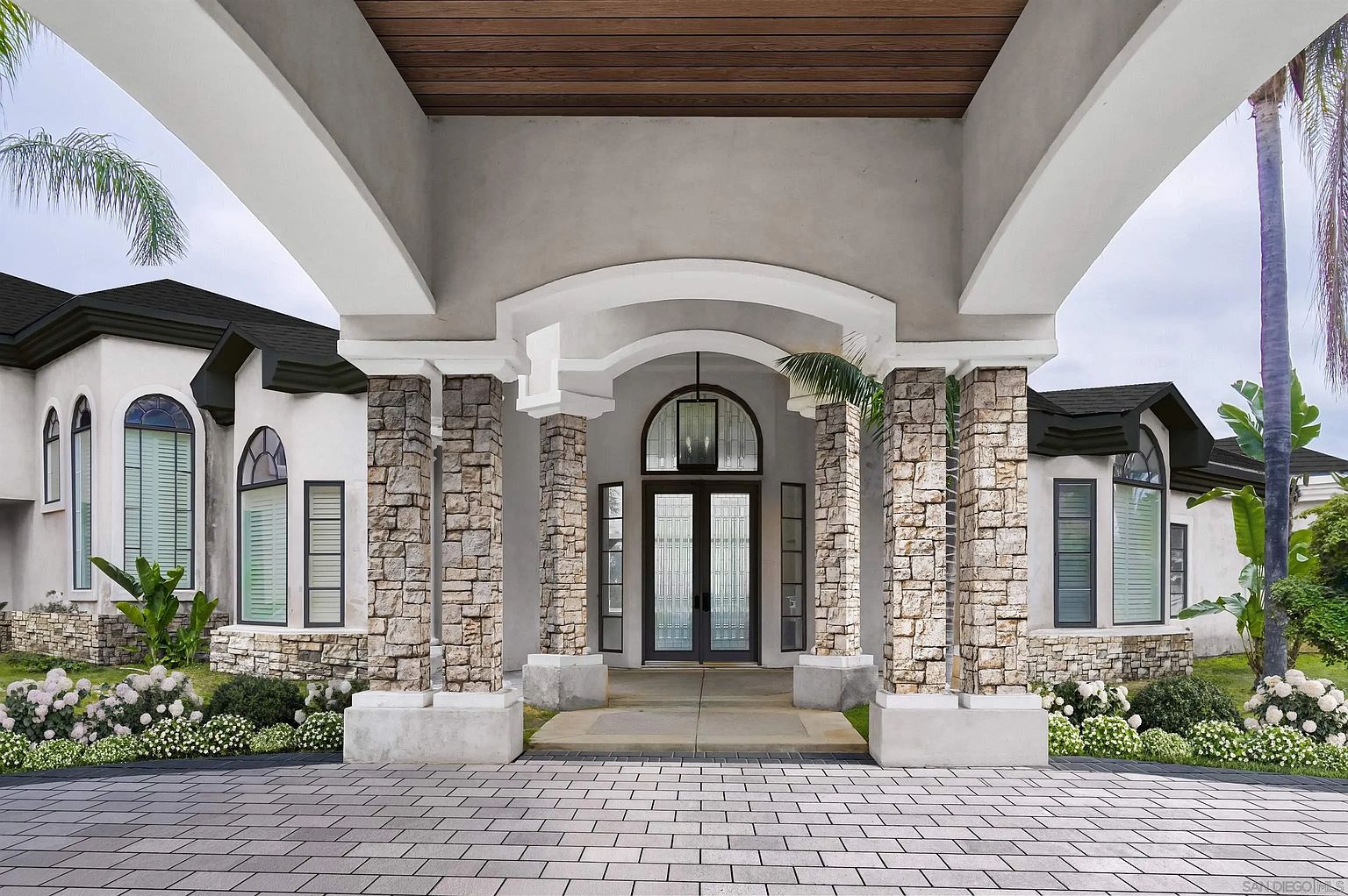 This is a grand entryway to a luxurious home, featuring stone-clad columns supporting a covered portico with a wooden ceiling. The double front doors are framed by sidelights and an arched transom window, creating an inviting entrance. The paved driveway and manicured landscaping enhance the property's curb appeal.
