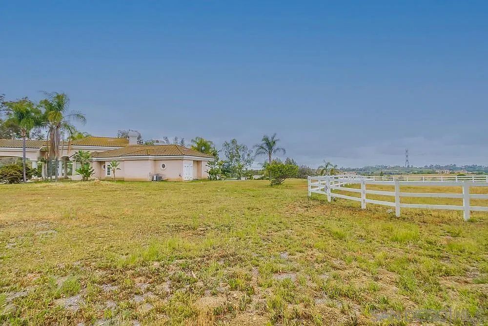The image showcases a sprawling yard with a well-maintained lawn, partially dry, leading up to a single-story house with a light-colored exterior and a tiled roof. A white wooden fence runs along the edge of the property, defining the boundary. The scene is bathed in natural light under a clear sky, creating a sense of openness and tranquility.