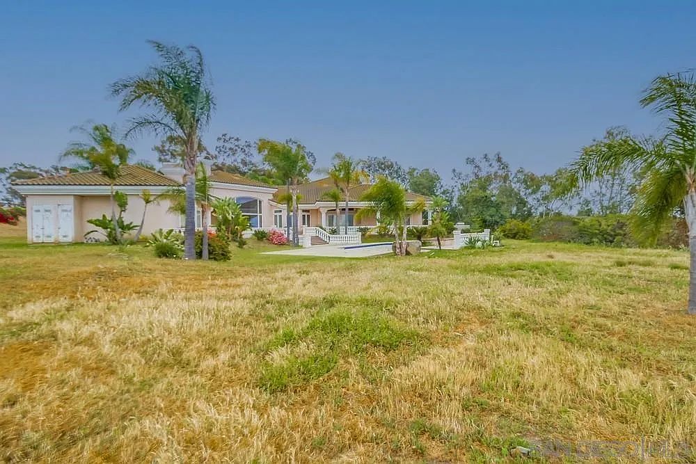 This image showcases the front exterior of a luxurious single-story home with a Mediterranean architectural style. The property features a well-manicured lawn, palm trees, and a swimming pool visible in the background. The house is painted in a light beige color, complemented by a tiled roof and elegant white columns.