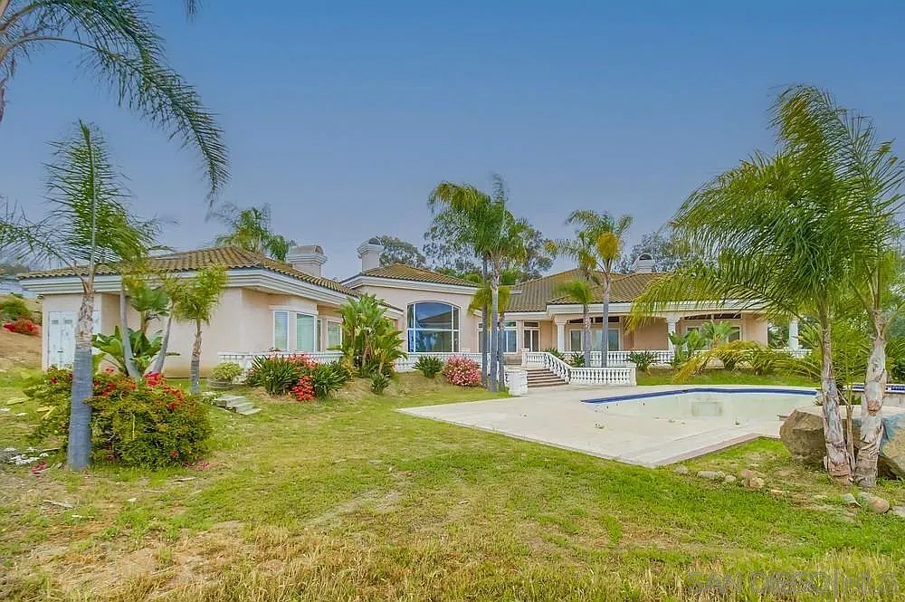 This image showcases the rear exterior of a luxurious home, featuring a well-manicured lawn, a swimming pool, and several palm trees. The house itself has a light-colored facade with a tiled roof, and the overall impression is one of upscale living and outdoor relaxation. The perspective is from ground level, providing a wide view of the property.