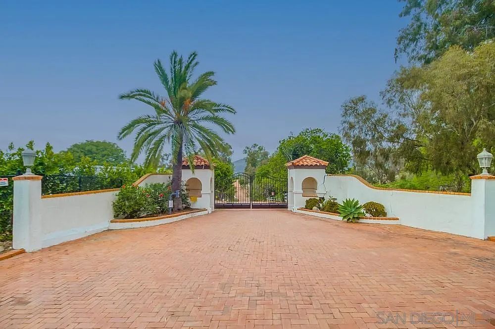 The image showcases a grand entryway featuring a brick-paved driveway leading to an ornate wrought-iron gate. Flanking the driveway are white stucco walls with arched niches topped with red tile roofs, adding a touch of Mediterranean style. A mature palm tree stands prominently on one side, enhancing the property's curb appeal and creating a welcoming impression.