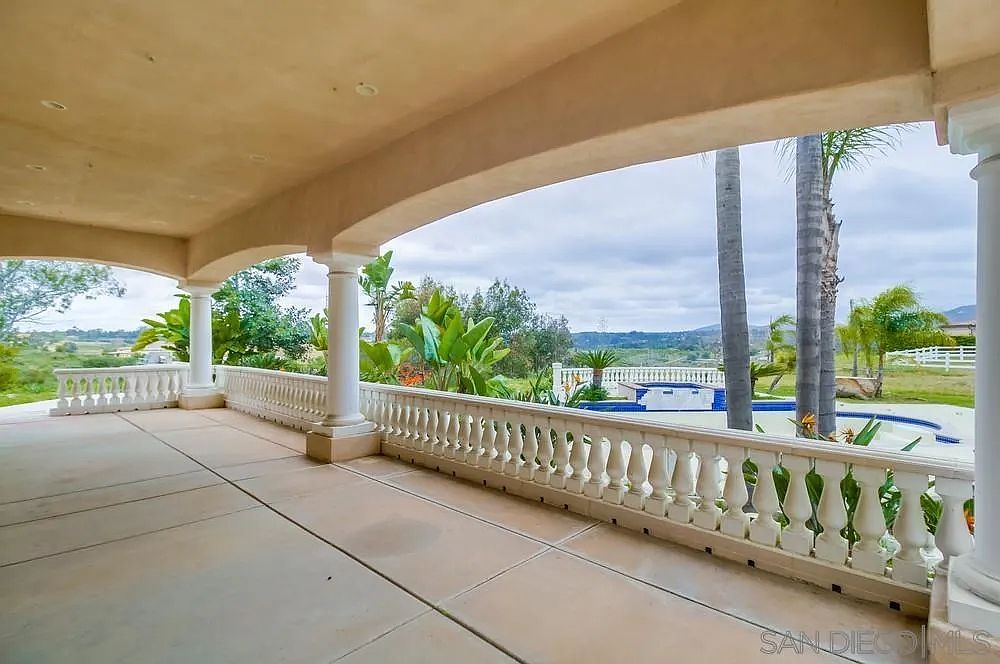 This image showcases a covered patio or balcony area with a decorative balustrade. The space features a concrete floor and is supported by columns. Beyond the patio, there is a view of a pool, landscaping, and a distant landscape, suggesting a luxurious outdoor living space.