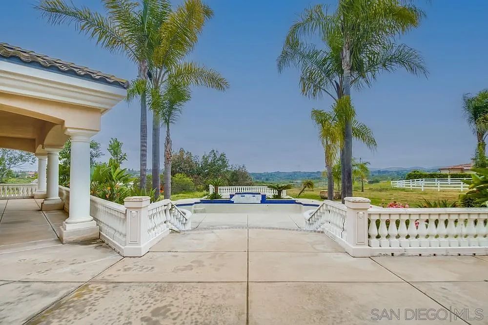 This image showcases an empty pool area with a decorative white balustrade surrounding it, flanked by palm trees and a view of the landscape beyond. The pool is the focal point, suggesting a luxurious outdoor living space. The architecture of the adjacent building adds to the upscale feel of the property.