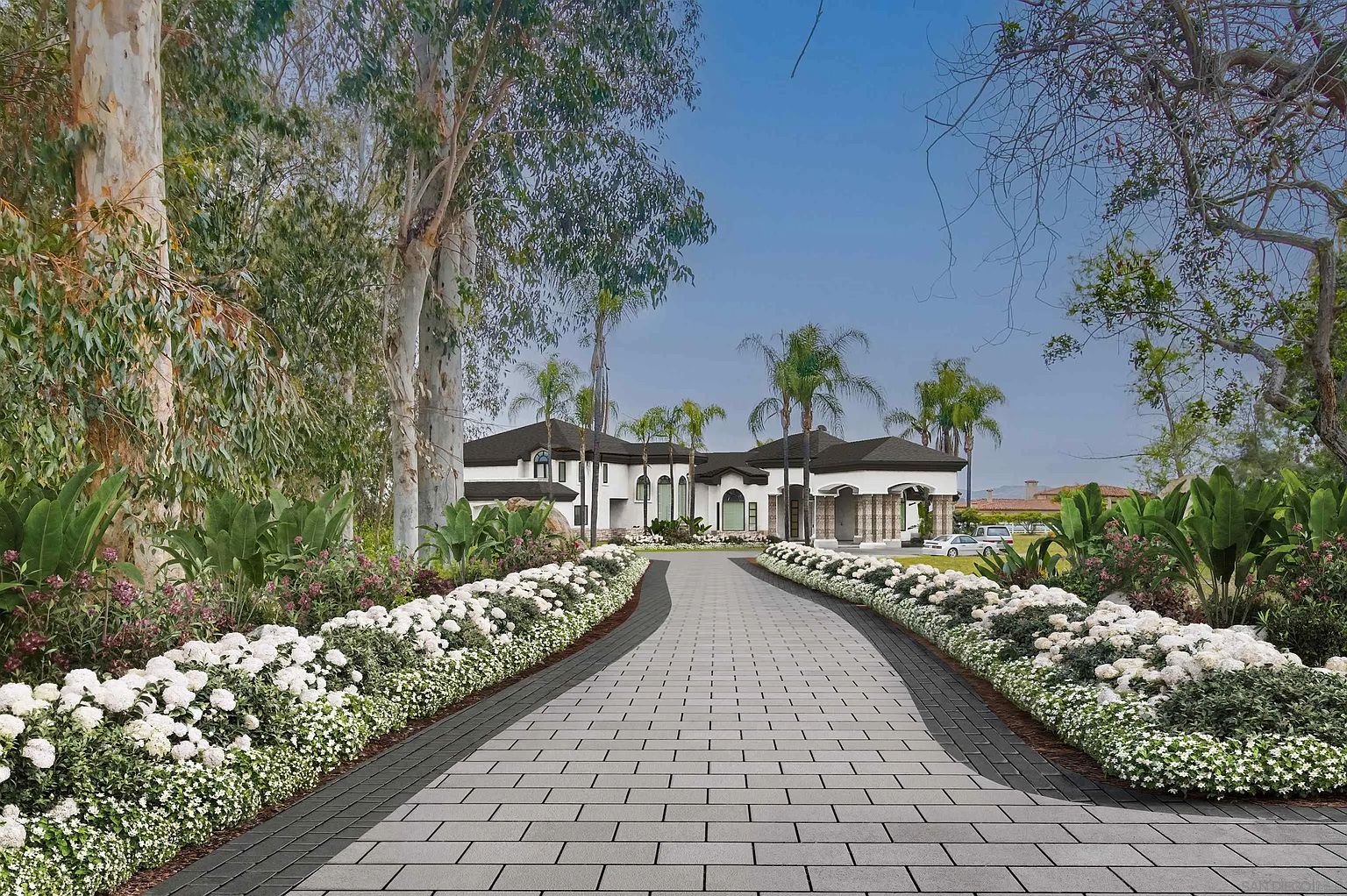 This image showcases the grand front view of a luxurious estate. A long, paved driveway lined with meticulously arranged white flowers leads to the impressive white facade of the house, which features a dark roof, arched entryways, and stately columns. Palm trees and lush greenery surround the property, creating a serene and upscale ambiance.