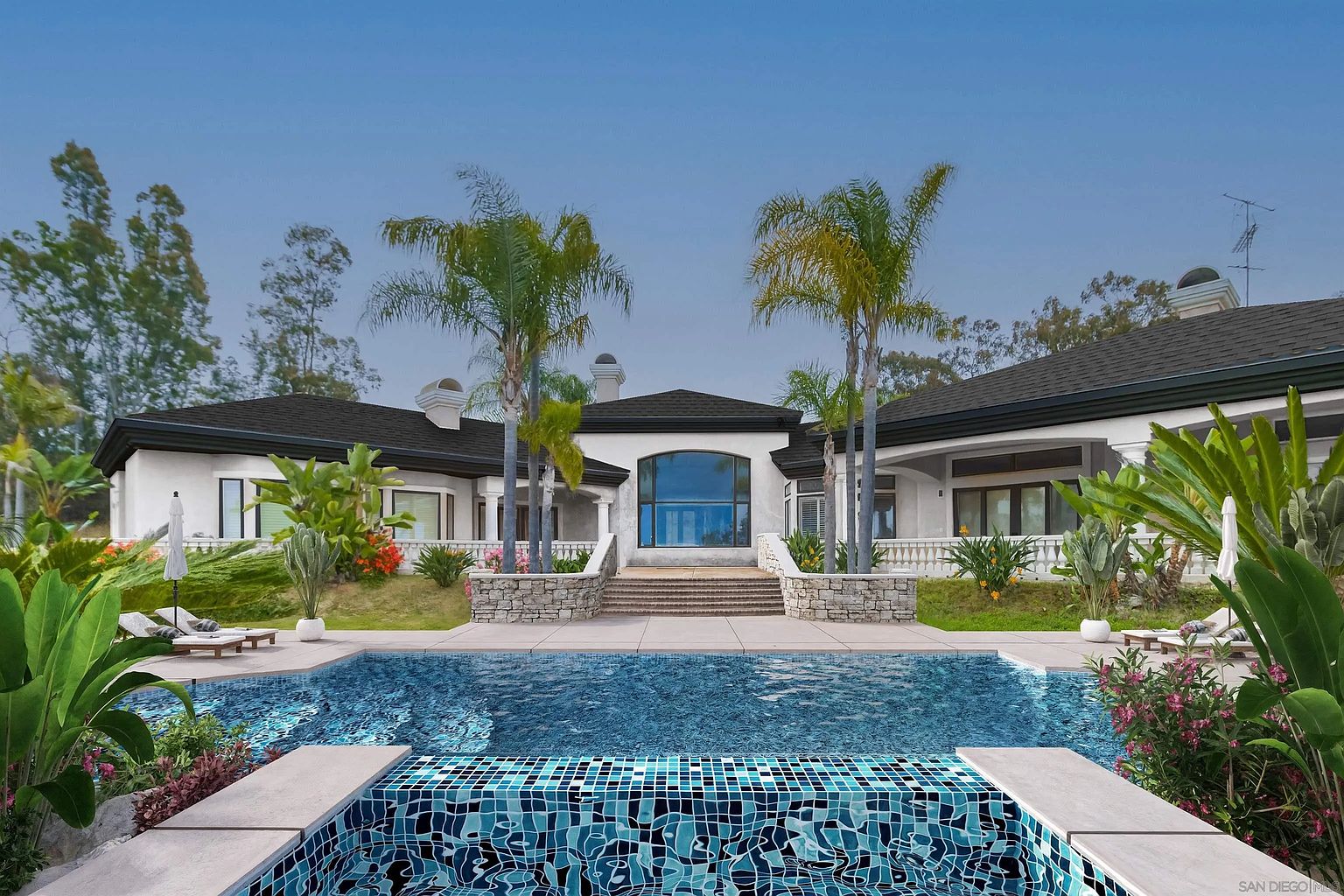 This image showcases a luxurious backyard with a stunning pool as the focal point. The pool features a unique tile design and is surrounded by lush landscaping, including palm trees and flowering plants. In the background, a grand house with a contemporary design and large windows adds to the overall impression of upscale living and relaxation.