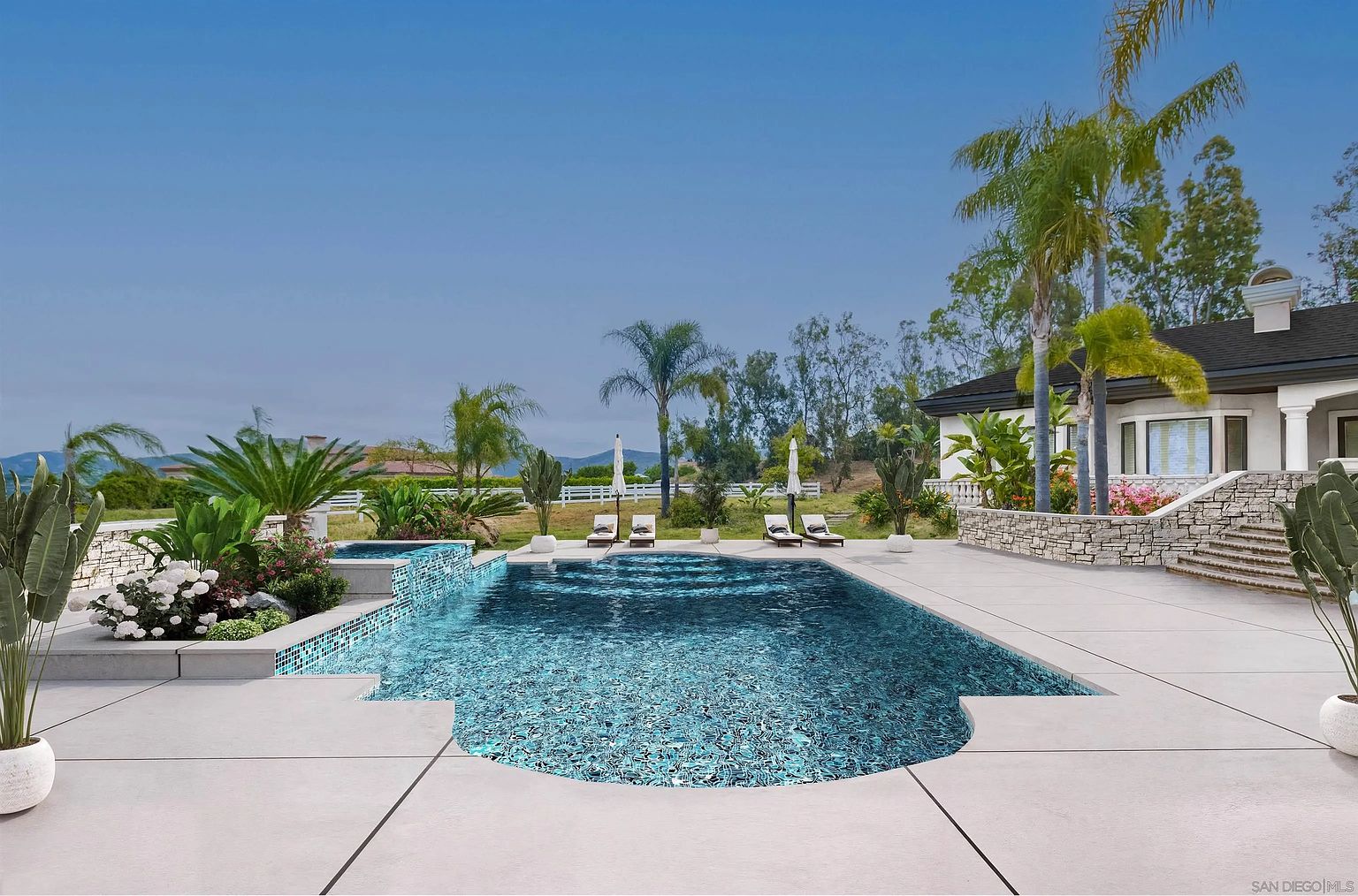 This image showcases a luxurious backyard pool and spa area. The pool features a unique shape and is surrounded by a spacious patio, lounge chairs, and lush landscaping, including palm trees. The property includes a well-maintained house in the background, suggesting a high-end residential setting.