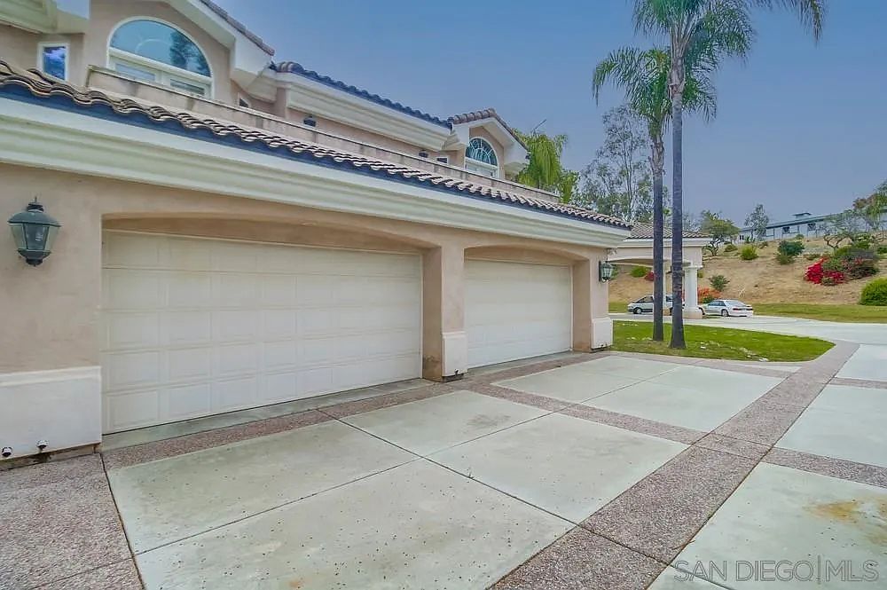 This image showcases the exterior of a home, focusing on the garage area. The garage features two white doors, complemented by a paved driveway with decorative stone accents. The house has a light beige exterior with a tiled roof, and a palm tree is visible in the background, adding to the property's curb appeal.
