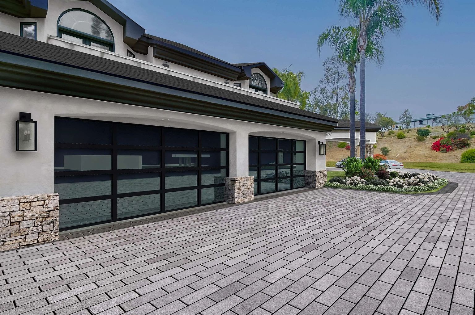 This image showcases a modern garage exterior with two black-framed glass garage doors, complemented by stone accents on the pillars. The driveway is paved with gray bricks, leading to a well-maintained lawn and landscaping with a palm tree in the background. The overall impression is one of contemporary elegance and curb appeal.