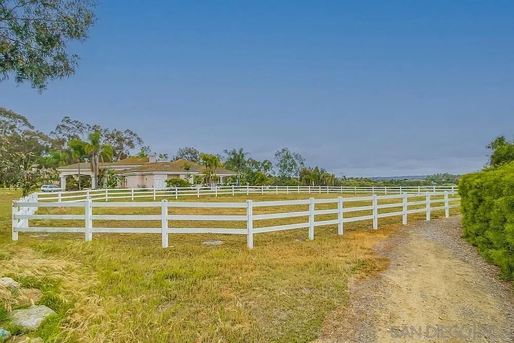 This exterior shot showcases a sprawling yard enclosed by a white fence, suggesting a spacious property. A well-maintained lawn and a glimpse of the house in the background create a serene and inviting atmosphere. A dirt road leads into the property, adding a rustic touch.