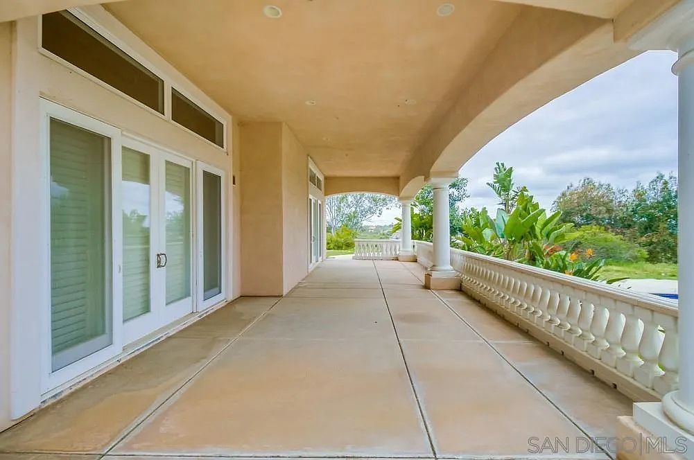 This image showcases a spacious outdoor patio or balcony area. The space features a covered walkway with architectural columns and a decorative balustrade, offering a blend of elegance and functionality. Large glass doors provide access to the interior, while the open side reveals a view of lush greenery and landscaping, enhancing the property's appeal.