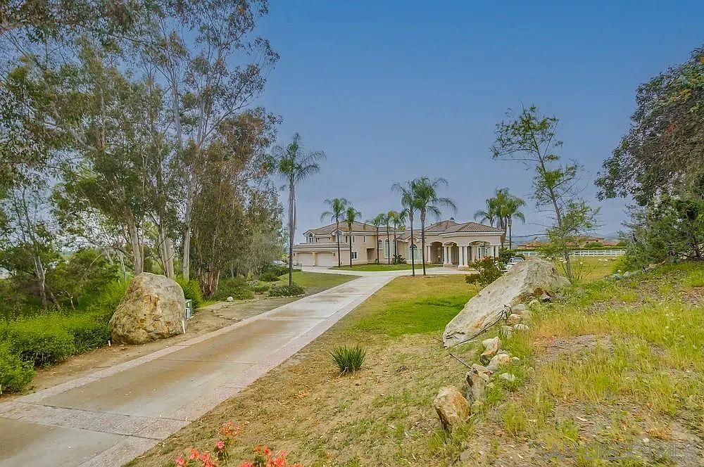 This image showcases the front exterior of a grand estate, featuring a long driveway leading to a large, luxurious home with a beige facade and a red tile roof. Palm trees and manicured lawns accentuate the property's elegance, while large boulders flank the driveway, adding a natural element to the landscape. The overall impression is one of opulence and privacy.