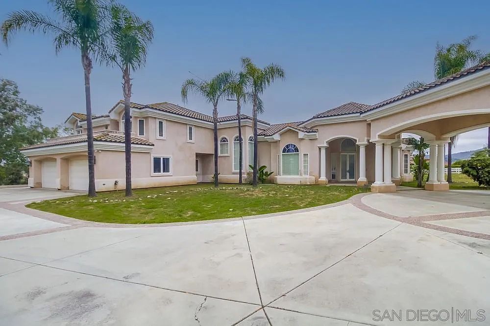 This is a front exterior view of a large, luxurious single-family home. The house features a light beige stucco exterior, a red tile roof, and a circular driveway. Palm trees are scattered around the front yard, adding to the property's curb appeal and upscale feel.