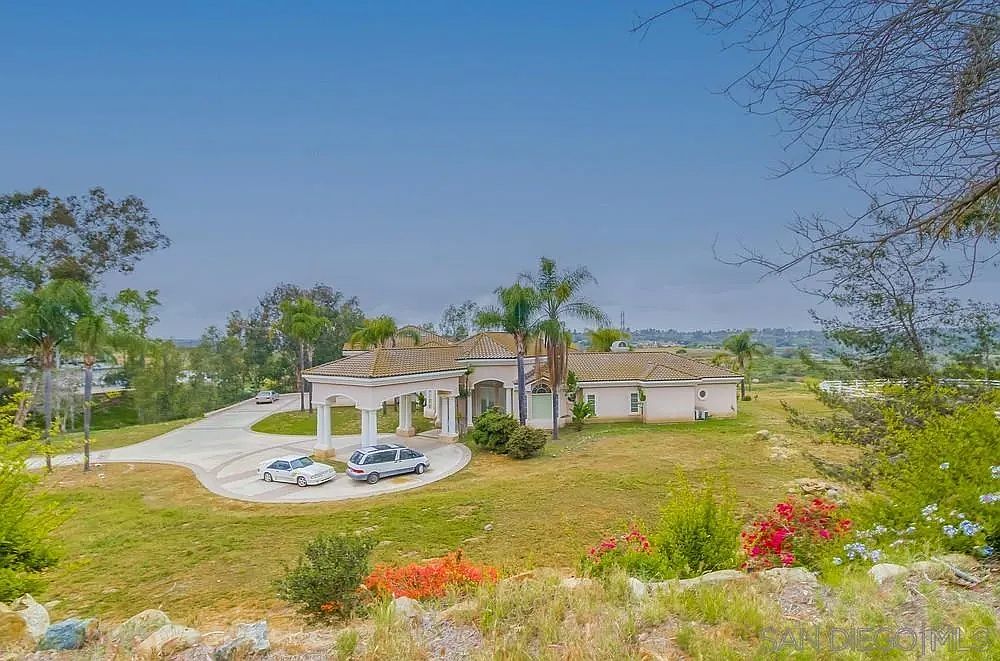 This aerial view showcases a sprawling estate with a circular driveway leading to a grand entrance. The property features lush green lawns, mature trees, and well-maintained landscaping. The house itself has a light-colored exterior with a tiled roof, exuding a sense of luxury and privacy.