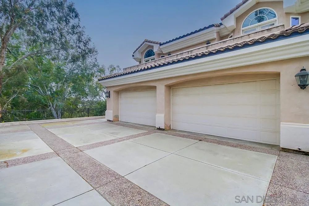 This image showcases a two-car garage of a residential property. The garage doors are white and appear well-maintained. The driveway is a combination of concrete slabs and decorative aggregate, adding visual interest to the exterior. The building's facade is light-colored with architectural details such as arched windows and a tiled roof, suggesting a Mediterranean or Spanish style.