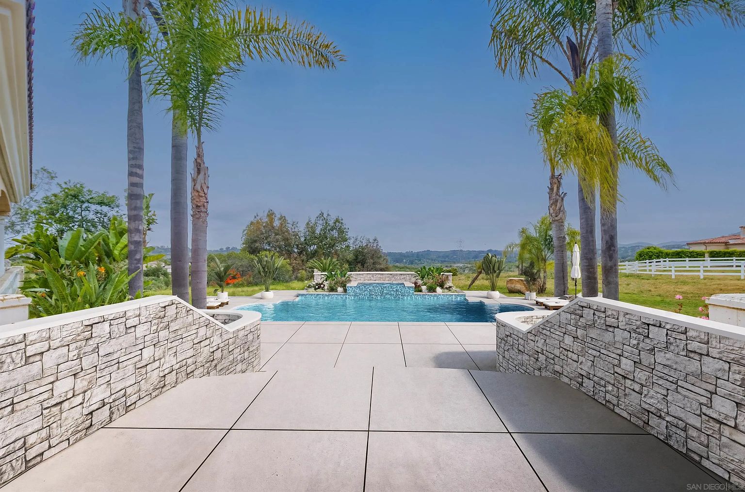 This image showcases a luxurious outdoor pool area, framed by tall palm trees and elegant stone retaining walls. The pool features a waterfall element, adding a touch of serenity and sophistication. The surrounding patio is spacious and well-maintained, suggesting a perfect space for relaxation and entertainment.