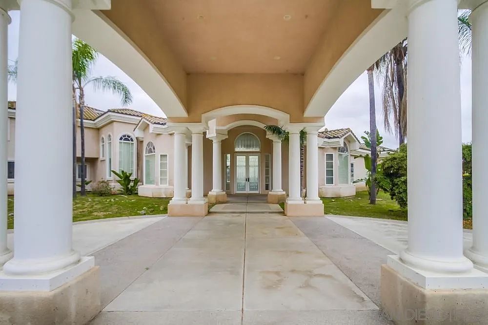 This image showcases the grand entryway of a luxurious home, featuring a covered driveway supported by elegant white columns. The entrance leads to double glass doors, hinting at a bright and welcoming interior. The overall impression is one of sophistication and upscale living, with well-maintained landscaping visible on either side.