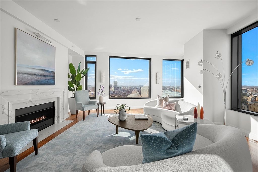 This is a bright and airy living room with a modern design. The room features large windows offering a city view, a fireplace with a marble surround, and contemporary furniture including curved sofas and armchairs. The color palette is primarily white and light blue, creating a serene and sophisticated atmosphere.