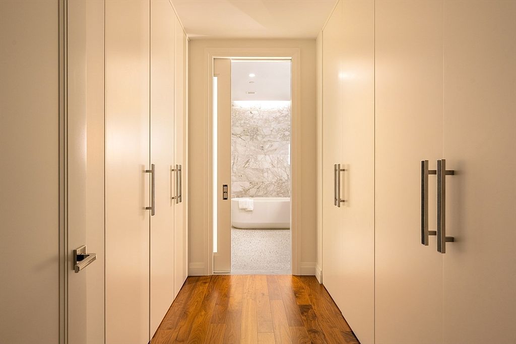 This interior shot showcases a hallway lined with built-in wardrobes or closets, leading to a bathroom. The warm wood flooring contrasts with the light-colored cabinetry, creating a welcoming atmosphere. The bathroom, visible through the doorway, features marble walls and a modern bathtub, suggesting a luxurious and well-appointed space.