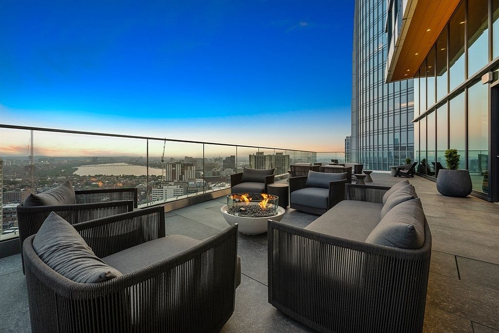 This image showcases a luxurious outdoor patio or balcony space, likely part of a high-rise apartment or condominium. The area features modern outdoor furniture, including comfortable seating arrangements around a stylish fire pit. A glass railing provides unobstructed views of the cityscape and skyline, enhancing the appeal of this outdoor living space.