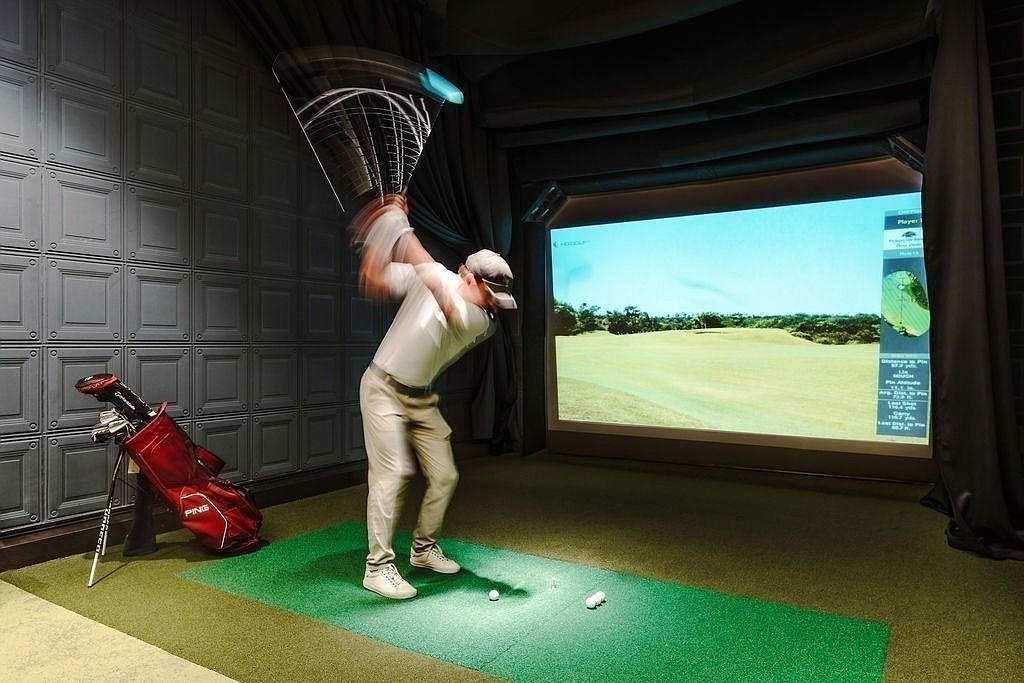 This image showcases an indoor golf simulator setup, featuring a golfer in motion against a screen displaying a virtual golf course. The room includes a golf bag, a green mat, and a padded wall, creating a dedicated space for practicing golf indoors. The overall impression is a high-tech and convenient amenity for golf enthusiasts.